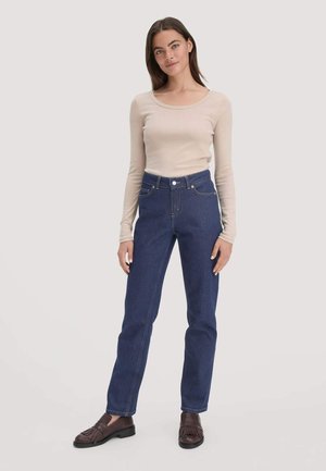 Vrouw staat in een beige lange mouwen top, hoge taille blauwe jeans en donkerbruine loafers tegen een effen lichte achtergrond.