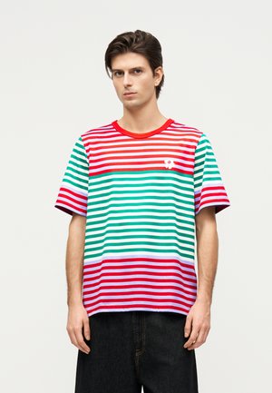 Fiatal férfi rövid ujjú, piros, zöld, fehér és lila csíkos pólóban, fekete nadrággal, sima háttér előtt állva.