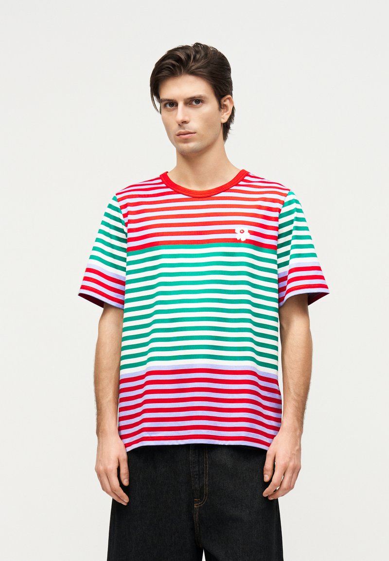 Fiatal férfi rövid ujjú, piros, zöld, fehér és lila csíkos pólóban, fekete nadrággal, sima háttér előtt állva.