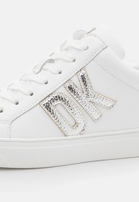Baskets blanches en matériau synthétique lisse, avec un logo "DK" argenté métallique sur le côté et un design classique à lacets.
