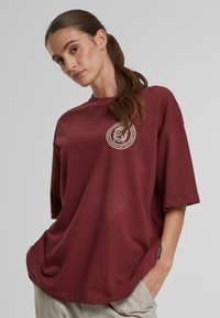 Burgundi oversized marškinėliai trumpomis rankovėmis, su apskritimo dizaino logotipu ant krūtinės, pagaminti iš minkštos medvilnės medžiagos.