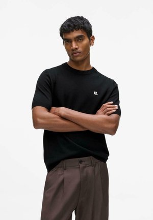 Ung mand stående med armene over kors iført en sort kortærmet skjorte og brune bukser mod en ensfarvet hvid baggrund.