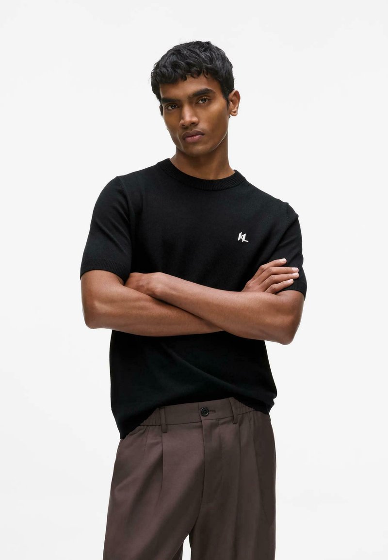 Jeune homme debout, bras croisés, portant une chemise noire à manches courtes et un pantalon marron sur un fond blanc uni.