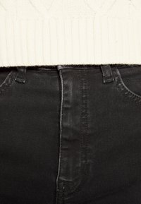 Jeans de denim preto com um acabamento suave, com cintura alta, bolsos frontais e costura preta, combinados com um suéter de malha creme.