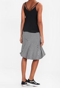 Donna in top senza maniche nero e gonna a campana a scacchi bianco e nero con sneakers nere, in piedi di spalle su sfondo bianco.