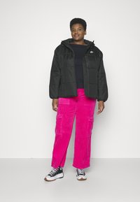 Veste noire rembourrée avec capuche, pantalon en velours côtelé rose vif à côtes, et baskets noires avec lacets blancs et semelles en caoutchouc.