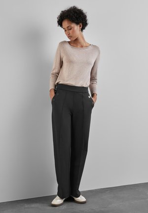 Donna con capelli ricci che indossa una maglia beige a maniche lunghe, pantaloni neri a vita alta e gamba larga con bottoni, e sneakers bianche, in piedi contro un muro grigio.