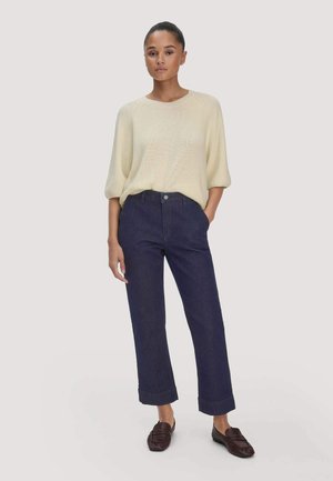 Vrouw draagt een crème kleurige gebreide trui, donkerblauwe cropped jeans en donkerbruine loafers, staand met één hand in de zak tegen een effen achtergrond.