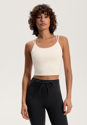 Helle beigefarbene, kurz geschnittene Tanktop mit dünnen Trägern und einer glatten Stoffoberfläche. Kombiniert mit schwarzen, hochgeschnittenen Hosen mit Kordelzug.