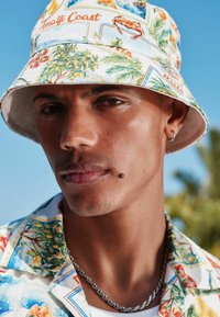 Chapeau seau avec un imprimé tropical comprenant des fleurs et des scènes côtières, porté avec une chemise à motifs ; chaîne en argent visible autour du cou.