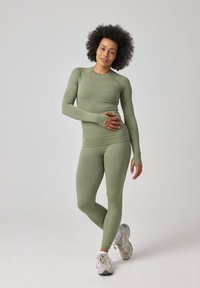 Olivengrünes, tailliertes Sportoberteil und Leggings aus dehnbarem Material, mit langen Ärmeln und Daumenlöchern, kombiniert mit hellen Sneakers.