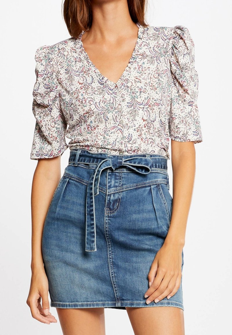 Femme portant un chemisier à manches bouffantes fleuri rentré dans une mini-jupe en jean bleue avec une ceinture nouée à la taille.