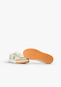Beige Leinenschuhe mit weißen Schnürsenkeln, grauen Akzenten und einer orangefarbenen Gummisohle. Besitzt ein Totenkopf-Design an der Seite und eine strukturierte Sohle.