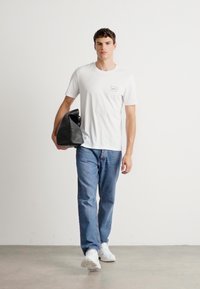 Ungdommelig mann som går, iført en hvit t-skjorte, blå jeans, hvite sneakers og bærer en svart lærveske i venstre hånd.