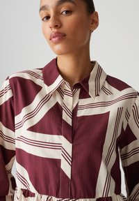 Vestito camicia bordeaux e crema con un colletto, caratterizzato da righe orizzontali e design geometrici, chiusura frontale con bottoni e vita arricciata.
