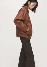 Brązowa skórzana kurtka o luźnym kroju, z dużymi kieszeniami i ściągaczem na dole, zestawiona z szerokimi ciemnymi spodniami.