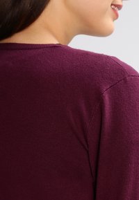 Vínový svetr s kulatým výstřihem a žebrovaným lemem, vyrobený z jemného pleteného materiálu s hladkou texturou. Zvýrazňuje oblast ramen.