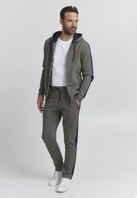 Grey zip-up hoodie with a navy lined hood and orange drawstrings, paired with matching sweatpants featuring side navy stripes and logo accents.