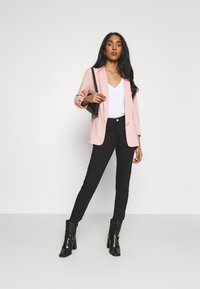 Blazer rosa claro sobre una camiseta blanca de escote en V, combinado con jeans skinny negros y botas negras de tobillo. El material parece suave y bien ajustado.