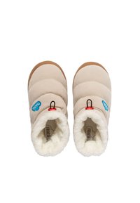 Botas de pantufla beige con parte superior acanalada, forro de piel sintética blanca, suelas de goma marrón y lengüetas rojas con un logo azul.