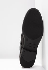 Semelle de chaussure noire avec un motif en caoutchouc texturé, présentant une forme arrondie et un petit talon. Contient une étiquette de taille "37" et un accent lisse.
