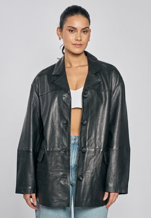 Vrouw met donker haar naar achteren gedragen draagt een zwart leren jack over een witte cropped top en lichtblauwe spijkerbroek tegen een grijze achtergrond.