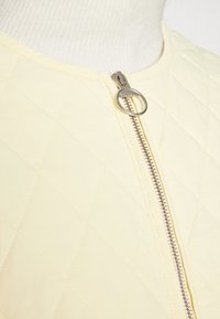 Gepolsterte gelbe Jacke mit rundem Kragen, ausgestattet mit einem silbernen Reißverschluss und einer glatten Textur. Das Muster besteht aus Rautenformen.