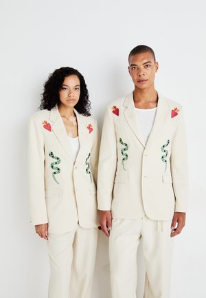 Two individuals wear cream blazers with intricate embroidered designs, featuring red hearts, green snakes, and decorative accents.
