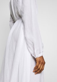 Biała sukienka z lnu z długimi, bufiastymi rękawami z zapięciami na guziki oraz talią wciętą. Miękka, teksturowana tkanina opada elegancko.