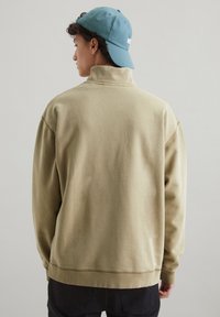 Jasno beżowy sweatshirt z wysokim kołnierzem, opadającymi ramionami i ściągaczami. Noszony z niebieską czapką, prezentując luźny i swobodny design.