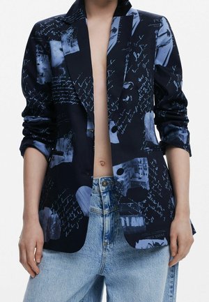 Person iført en mørkeblå blazer med lyseblå håndskrevet tekst og abstrakte designs, kombineret med lyseblå denimjeans.