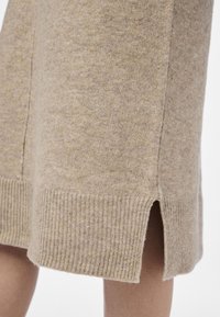Jupe en mélange de laine beige, avec un ourlet côtelé tricoté et des fentes latérales pour faciliter les mouvements. Texture douce avec une finition lisse.