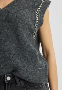 Dark gray knitted sleeveless top with studded rhinestone decoration near shoulder and black denim jeans waist visible.