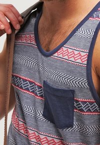 Débardeur sans manches en tissu bleu et gris avec des motifs géométriques agrémentés d'accents rouges et blancs, et une poche unie bleu marine sur le devant.