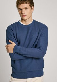 Maglione lavorato a maglia blu con scollo rotondo e polsini a coste. Presenta un logo discreto sul petto. Indossato sopra una camicia bianca.