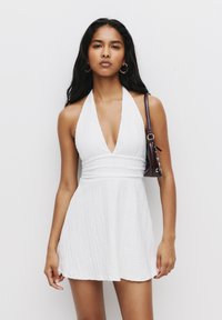 Weißes Halterkleid mit tiefem V-Ausschnitt, strukturiertem Stoff und taillierter Passform. Das Modell hält eine kleine bordeauxfarbene Handtasche mit dekorativen Akzenten.