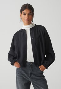 Zwarte jassen met knoopsluiting, met een ontspannen pasvorm, lichtgewicht stof en elastische manchetten, gedragen over een witte coltrui met donkere jeans.