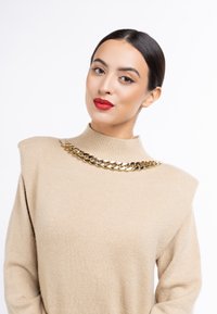 Pulover bej cu gât înalt și cu umăr padding, având un colier din lanț aurit. Textură netedă și design minimalist.