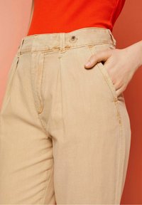 Gros plan d'une personne portant un pantalon beige avec une ceinture boutonnée et un haut rouge, main reposant dans la poche droite sur fond orange.