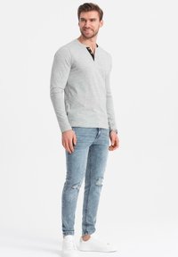 Grå långärmad henley-tröja med svarta accenter vid kragen, tillsammans med slitna ljusblå jeans och vita sneakers.