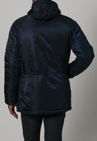 Navy blue insulated jacket with a hood, smooth satin-like finish, elastic waistband, and raglan sleeves. Features a gathered texture at seams.