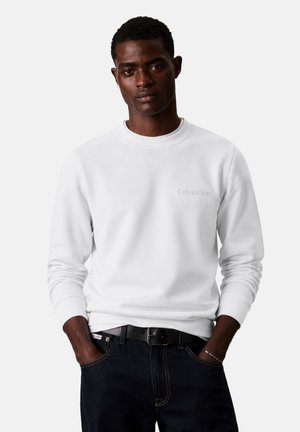 Modelo masculino con sudadera blanca Calvin Klein con las manos en los bolsillos de jeans oscuros frente a un fondo liso y claro.