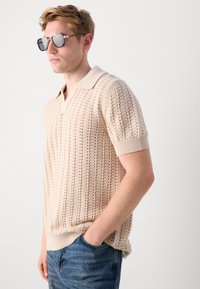 Beige knitted polo shirt with a textured, open pattern; short sleeves and a collared neckline paired with blue denim jeans.