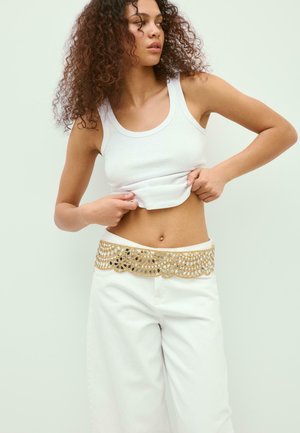 Donna con capelli ricci alza una canottiera bianca, indossa pantaloni bianchi con una larga cintura decorativa dorata, in piedi davanti a uno sfondo chiaro e semplice.