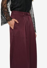 Burgundy high-waisted wide-leg trousers with pleats, featuring a button closure and a smooth texture. Black lace sleeve top in background.