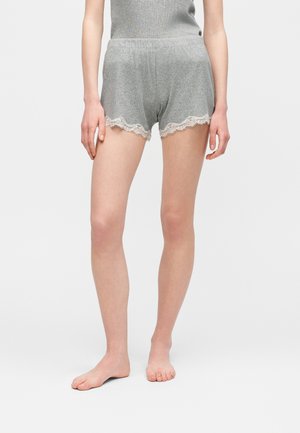 Kobieta ubrana w szare prążkowane szorty z białą koronkową lamówką, stojąca boso na jednolitym jasnym tle.