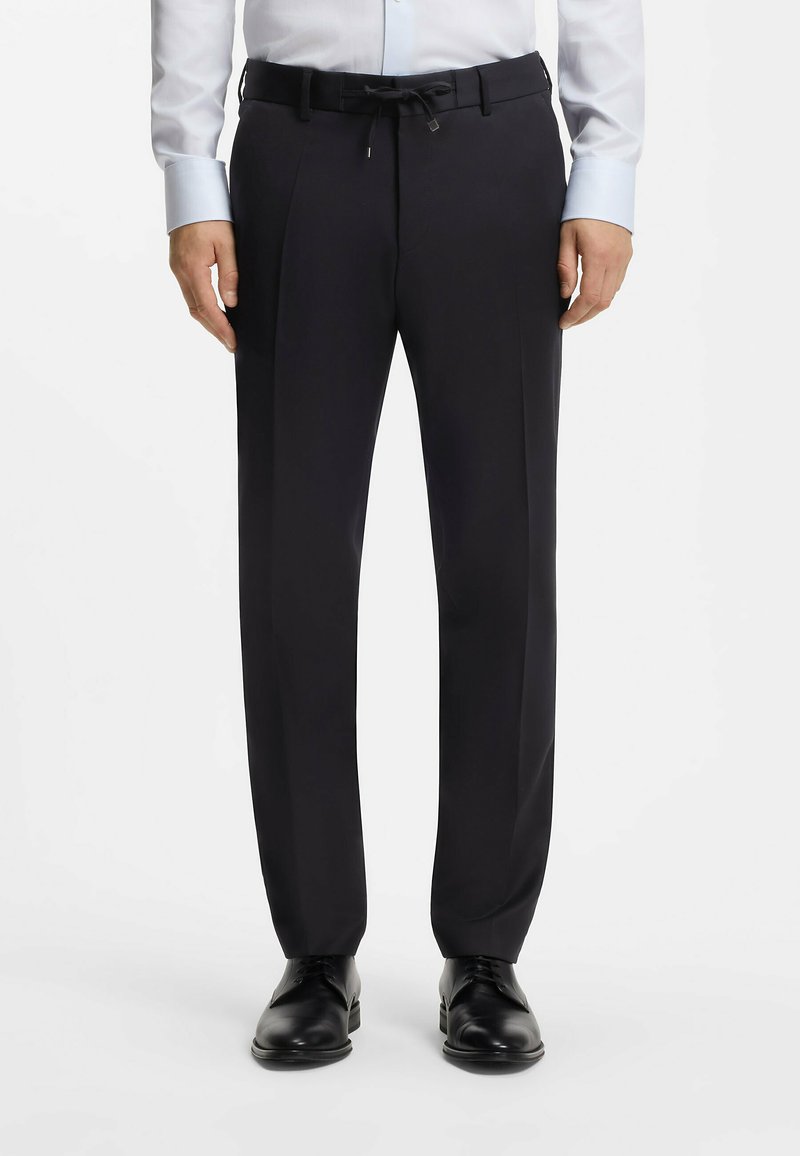 Homme portant un pantalon de costume noir à taille avec cordon, une chemise habillée bleu clair, une ceinture noire et des chaussures de ville noires, debout bien droit.