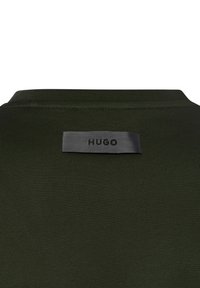 Dunkelgrüner Stoff mit einem kleinen grauen rechteckigen Etikett, das nahe der Kragennaht angenäht ist und den Text "HUGO" in schwarzen Großbuchstaben zeigt.