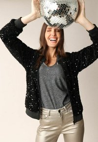 Veste bomber noire ornée de sequins avec des poignets côtelés, associée à un haut gris et un pantalon doré brillant. Le mannequin tient une boule à facettes miroir au-dessus de sa tête.