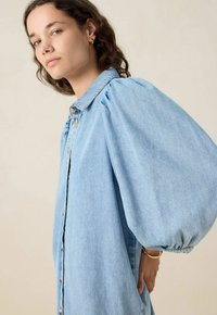 Jeune femme aux cheveux ondulés portant une robe en denim bleu clair avec des manches bouffantes volumineuses, ainsi que des boucles d'oreilles en or et un bracelet.